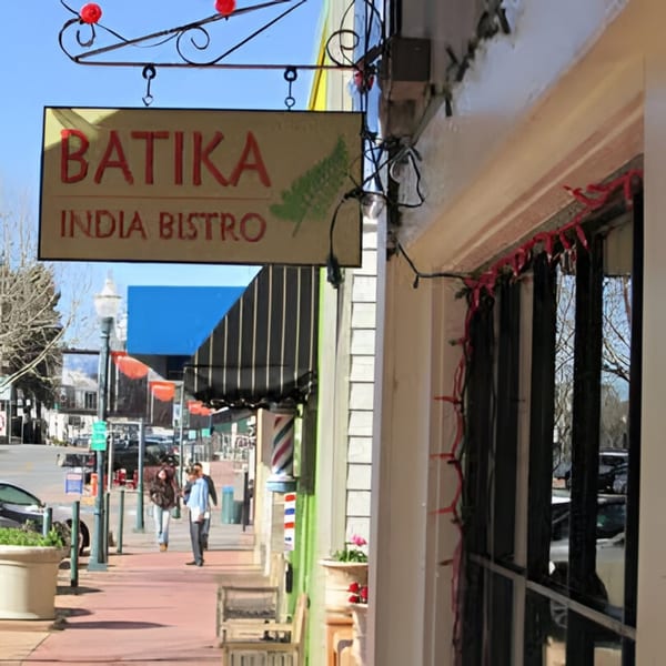 Batika India Bistro sign hanging above a sidewalk with people walking and storefronts lining the street.