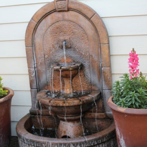 A rustic tiered water fountain with flowing water, flanked by potted plants with pink flowers.