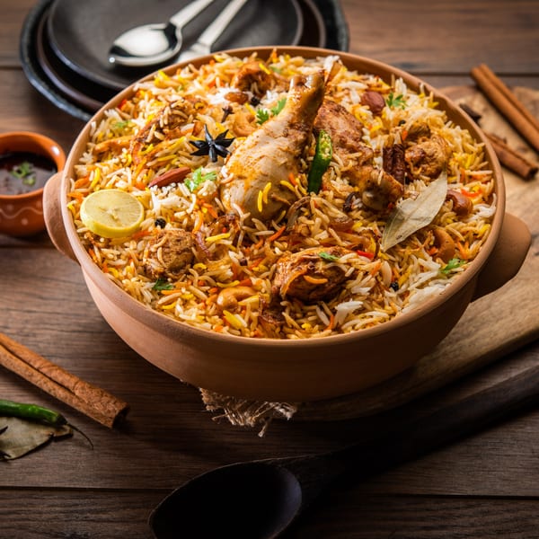 A large clay pot of Batika India Bistro's chicken biryani, garnished with lemon, spices, and herbs.