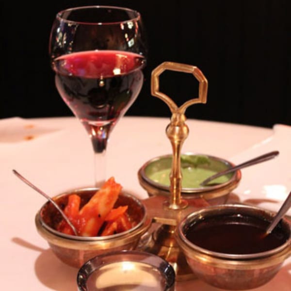 Red wine glass and Indian chutneys in small brass bowls on a white table.